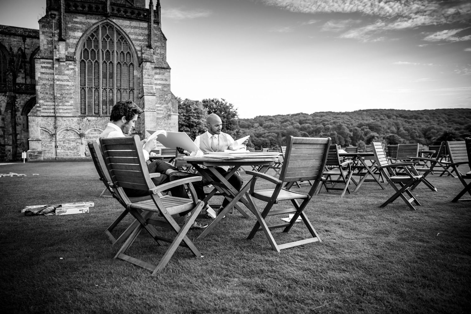 Black and white panoramic view of empty folding chairs set up on a lawn with gothic architecture.