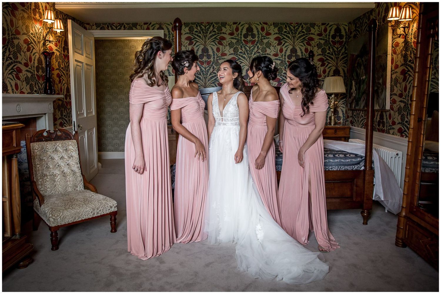 Bridesmaids in pink dresses pose with bride in elegant room with chandelier and decorative walls.