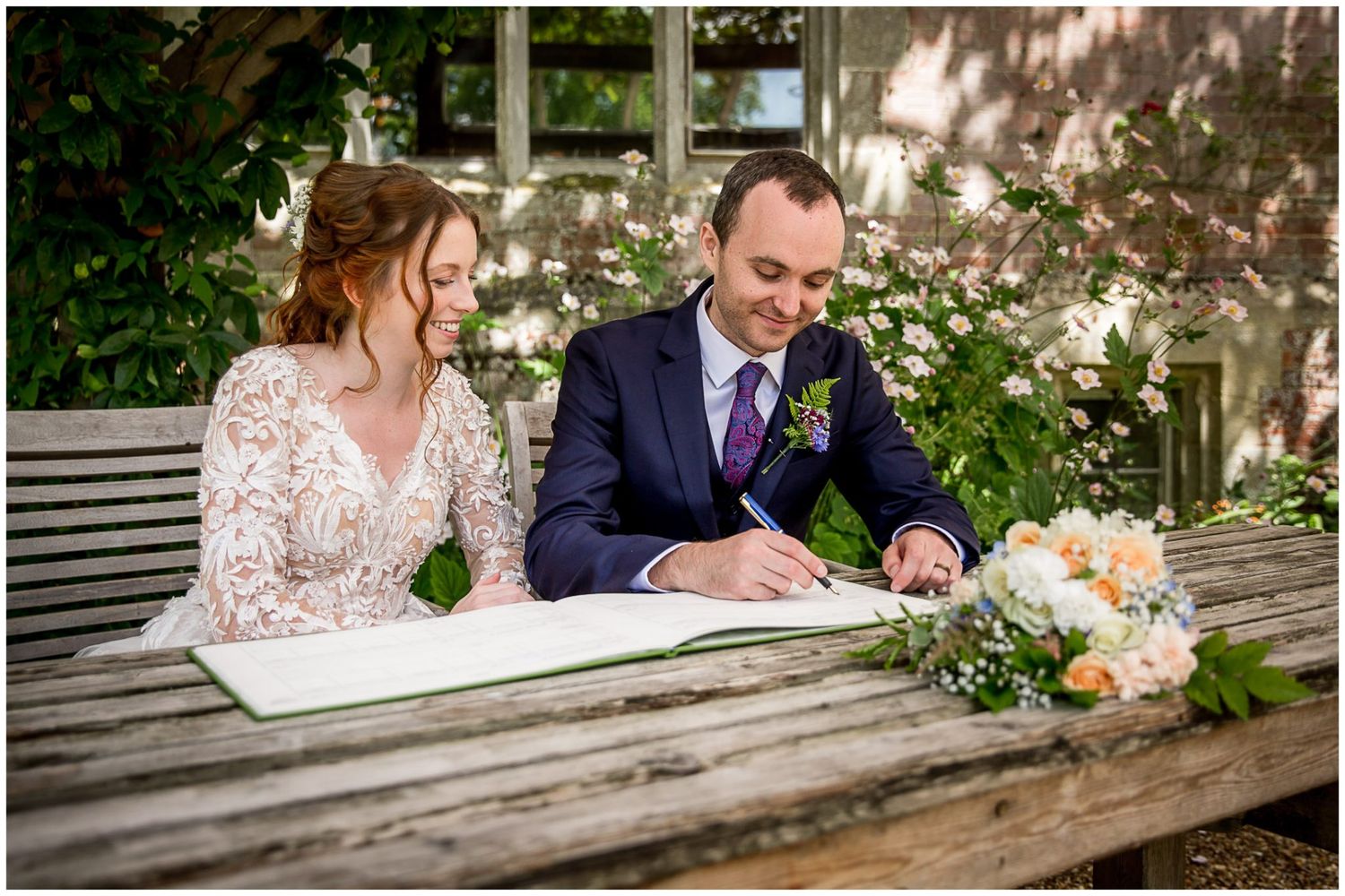 People signing documents at a rustic wooden table decorated with flowers during an outdoor celebration.