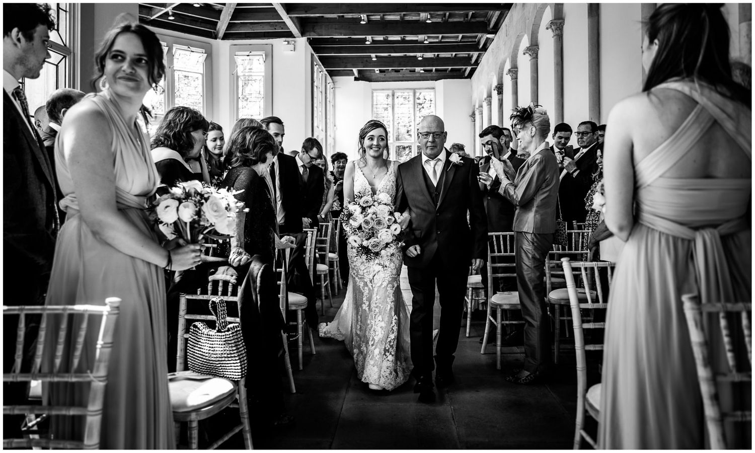 Wedding ceremony processional in sunlit hallway with white columns and decorative greenery.