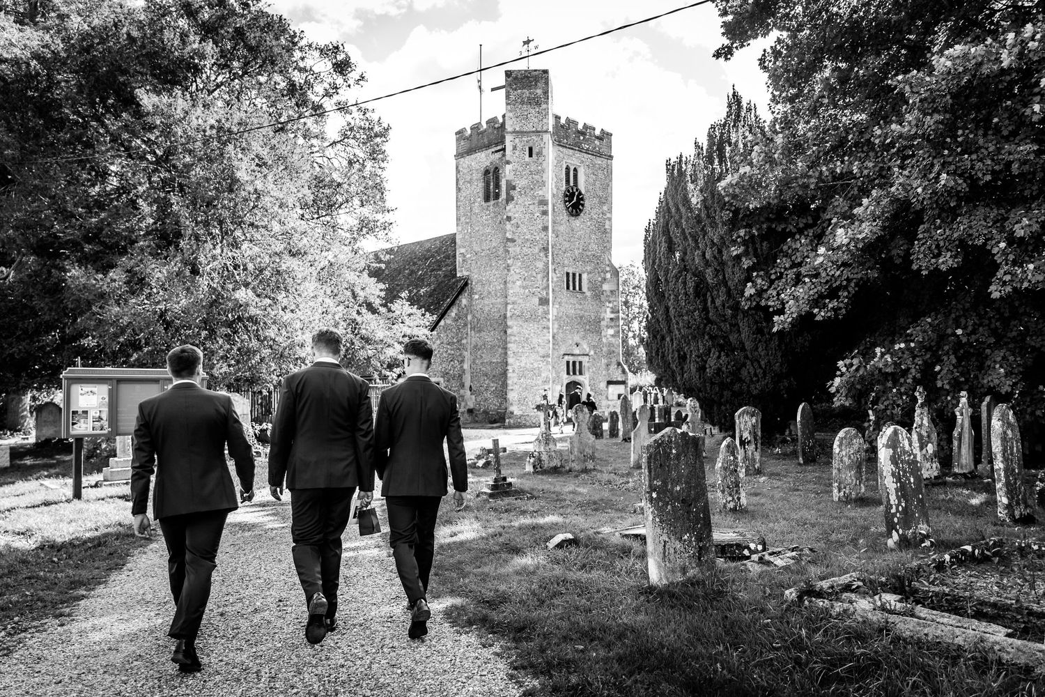Wedding party members walk through a scenic church graveyard with stone walls and trees.