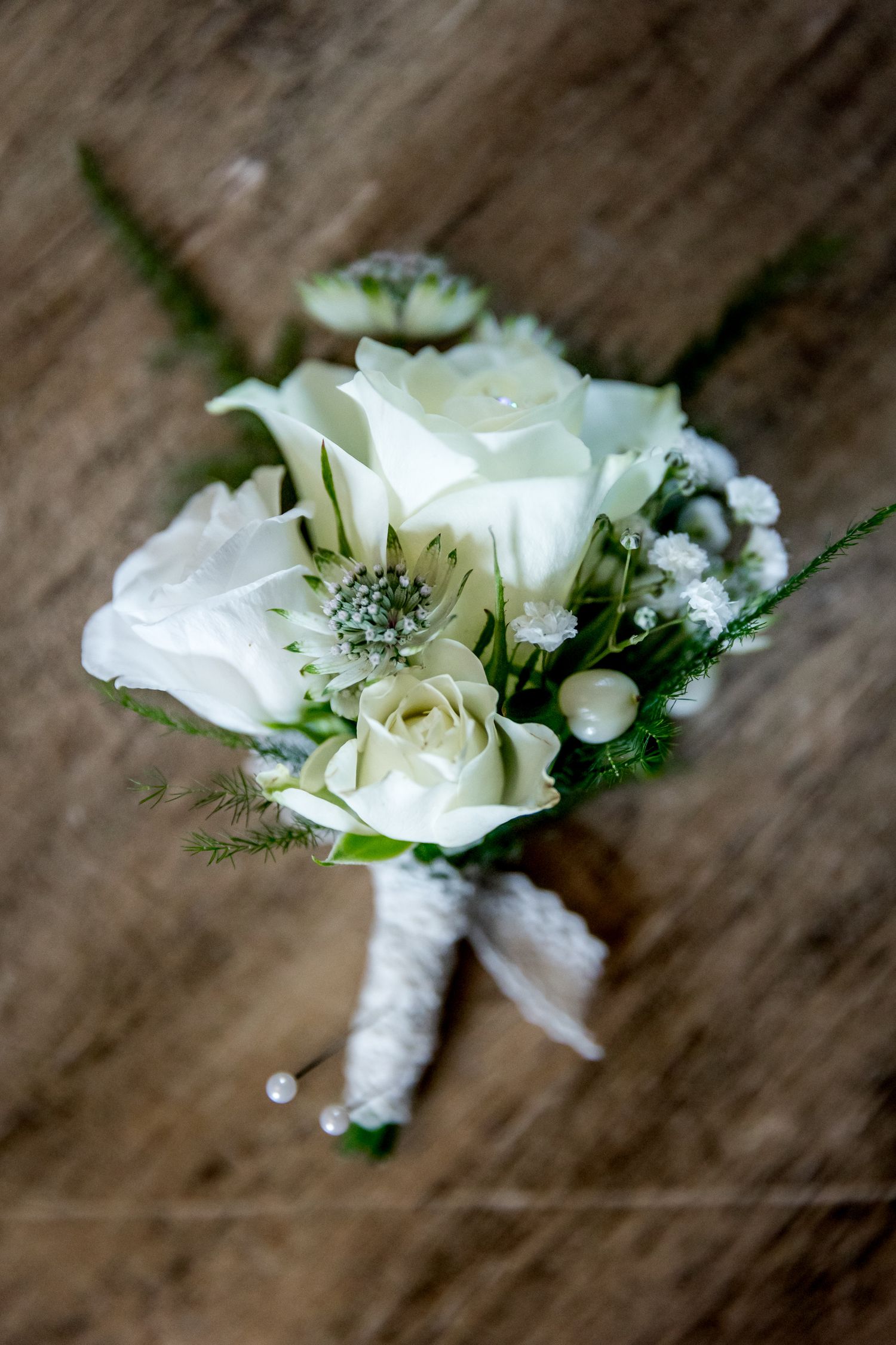 White rose boutonnieres with greenery and ribbon wrapping lined up on wooden surface.
