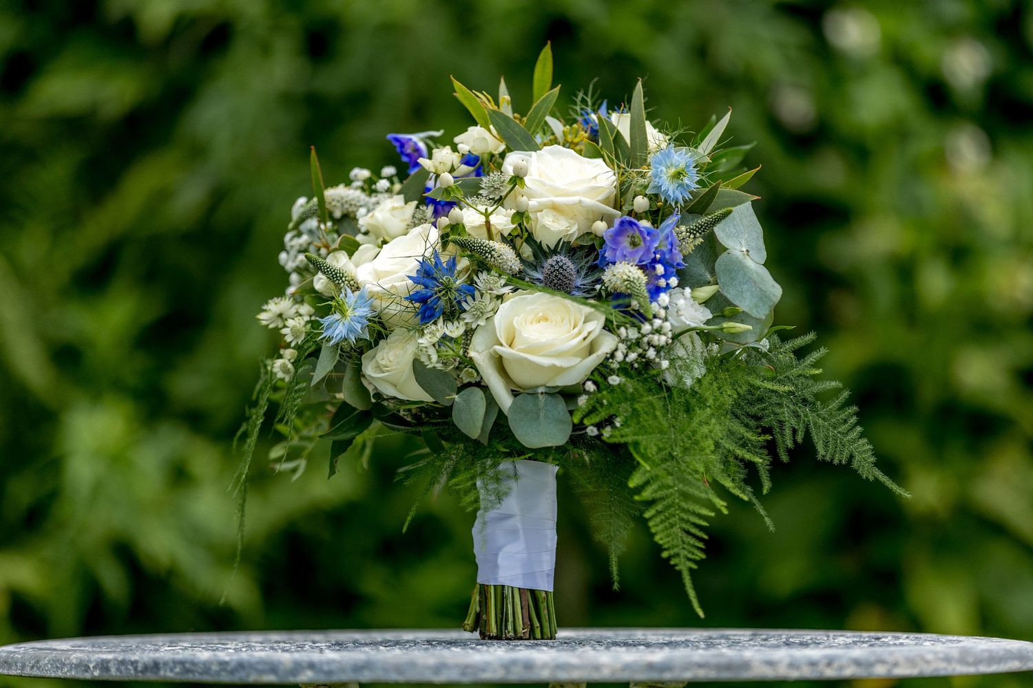 Wedding bouquet featuring white roses, blue flowers and green ferns against natural backdrop.
