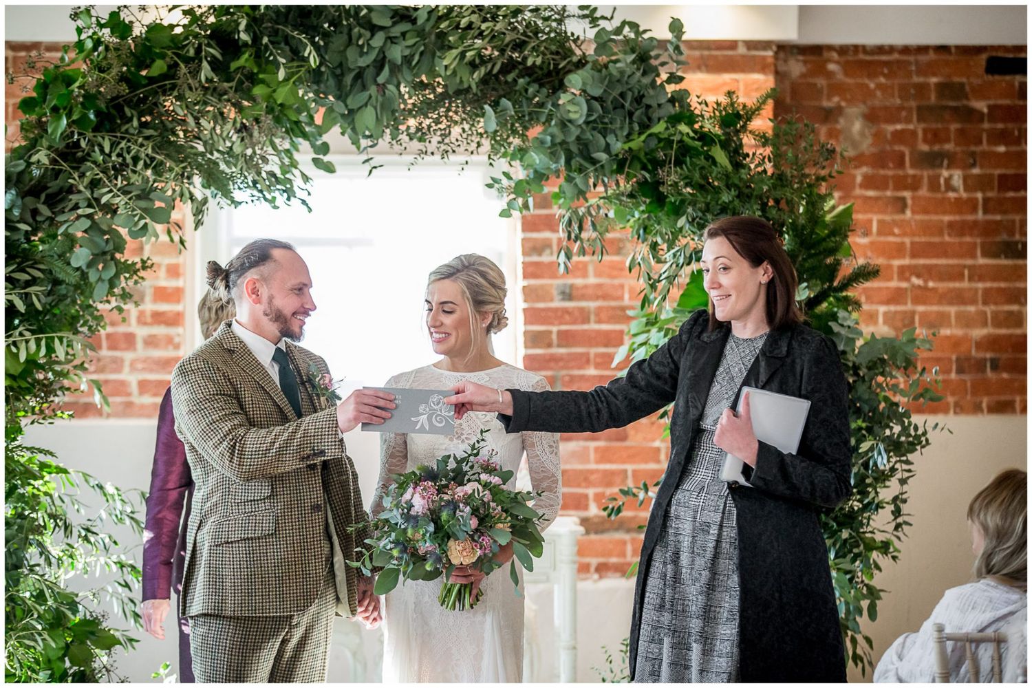 Wedding ceremony under a lush greenery arch with exposed brick wall backdrop.