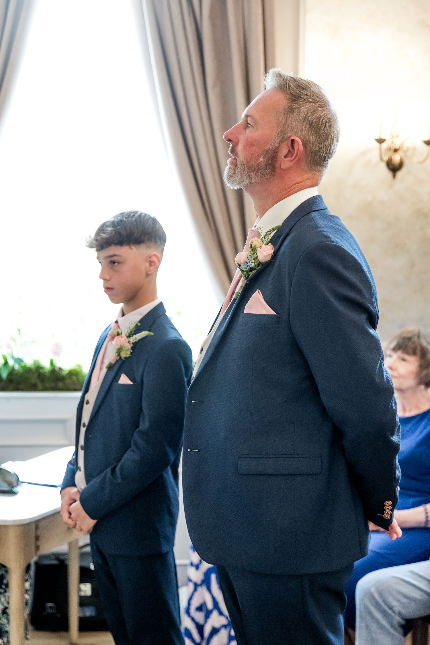 Wedding ceremony attendees in formal attire standing in sunlit room with curtains.