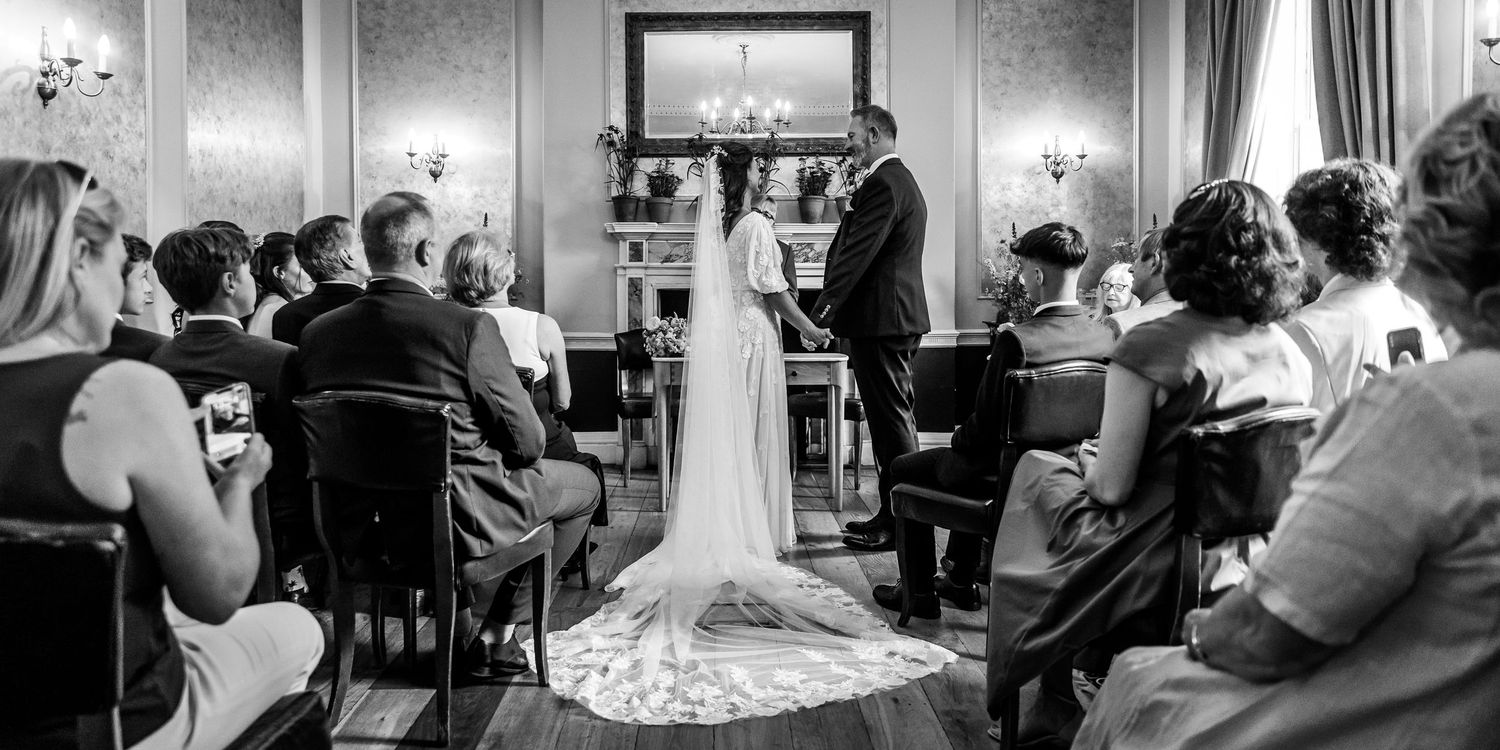 Black and white wedding ceremony photo in elegant room with guests seated.