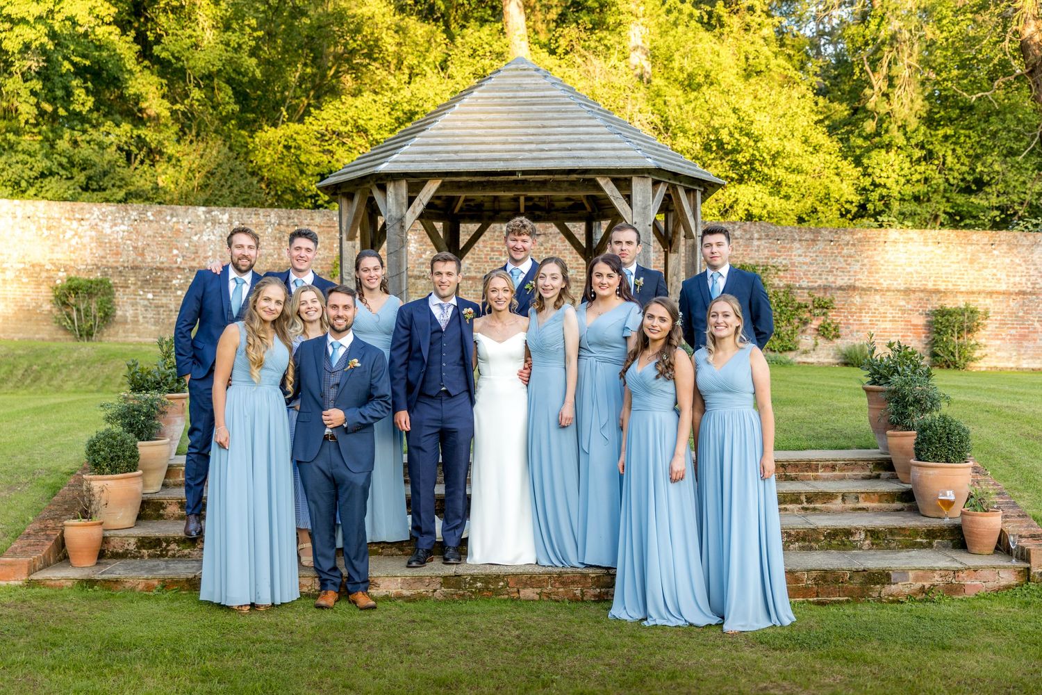 Wedding party in light blue bridesmaid dresses and navy suits pose together in front of a garden gazebo.