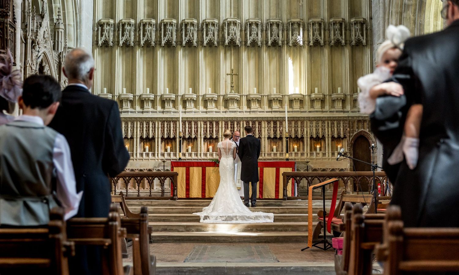 Wedding ceremony guests sit in ornate wooden church pews with Gothic architectural details visible in the background.