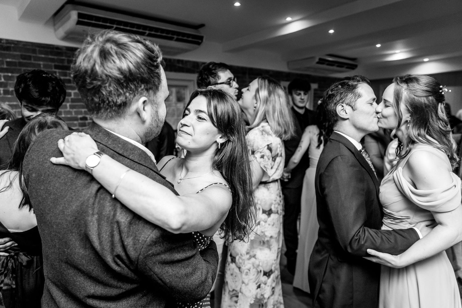 Black and white photo of wedding guests dancing in pairs at an indoor reception.