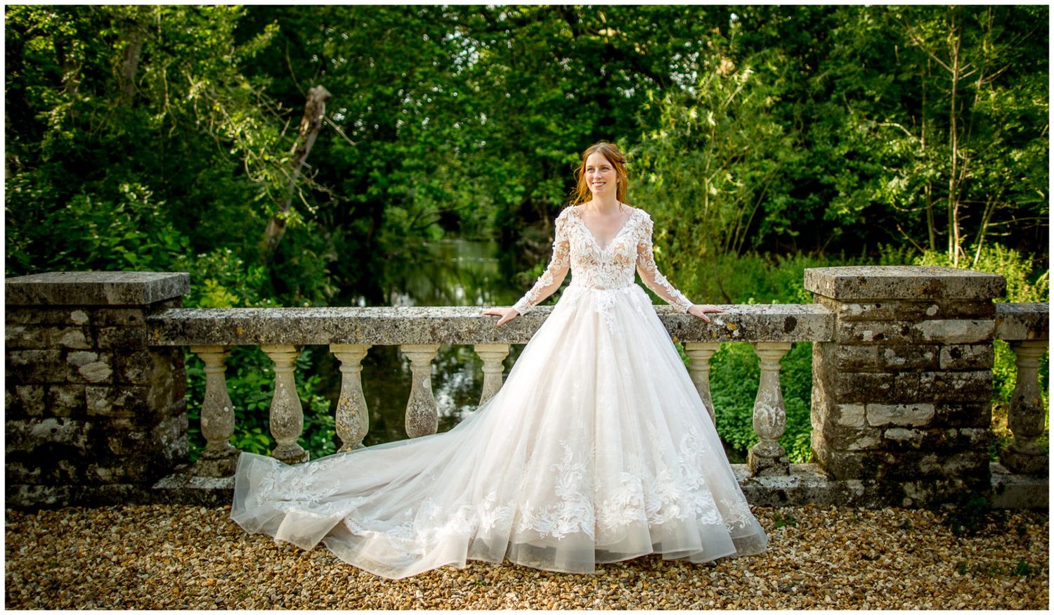 Bride in flowing white gown poses gracefully on stone bridge surrounded by lush garden scenery.