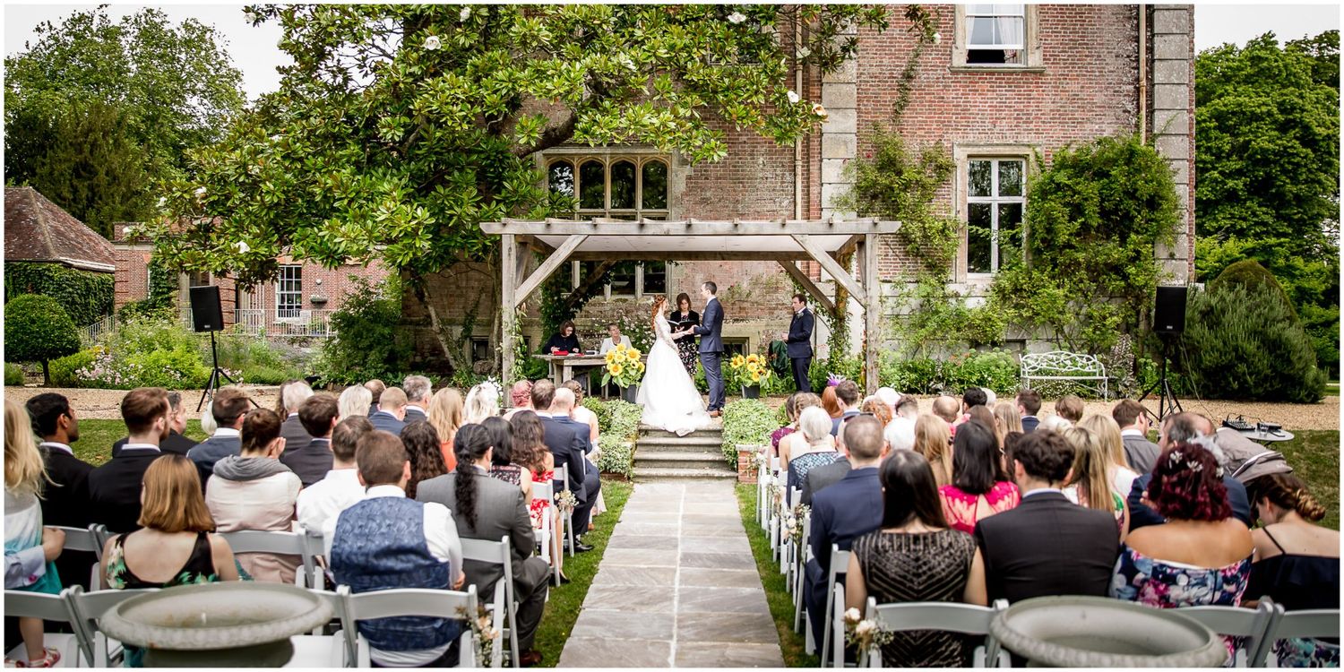 A group of wedding guests seated outdoors in rows of white chairs for a garden ceremony at a stone venue.