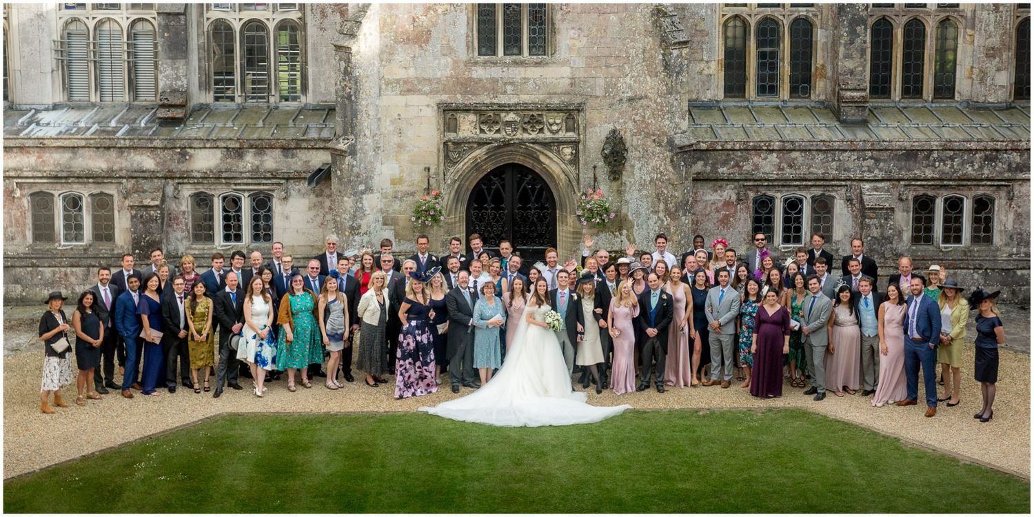 Large wedding party group photo taken outside a historic stone castle or manor building with Gothic architecture.