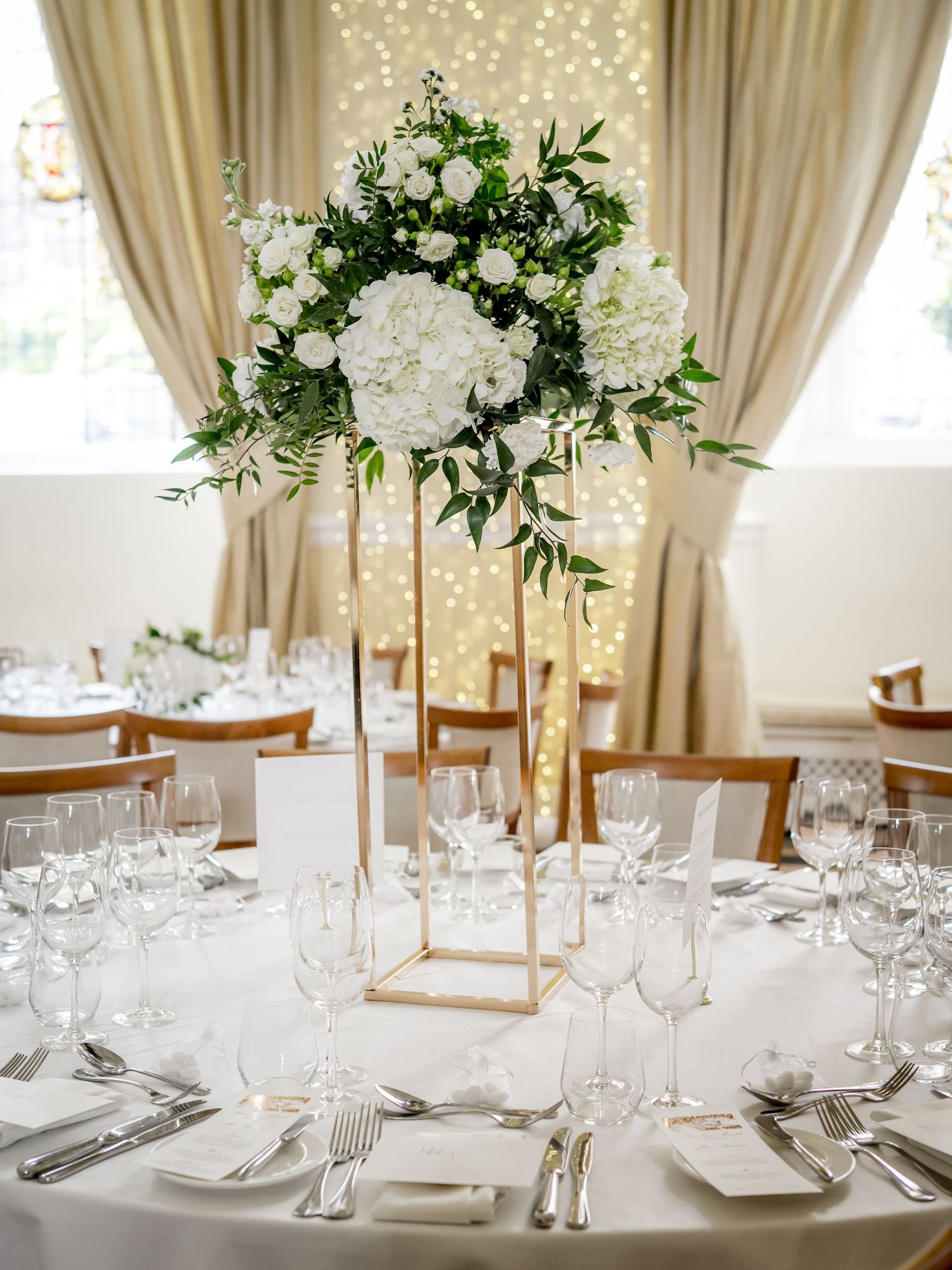 White floral centerpieces and crystal glassware adorn wedding reception tables with string light backdrop.