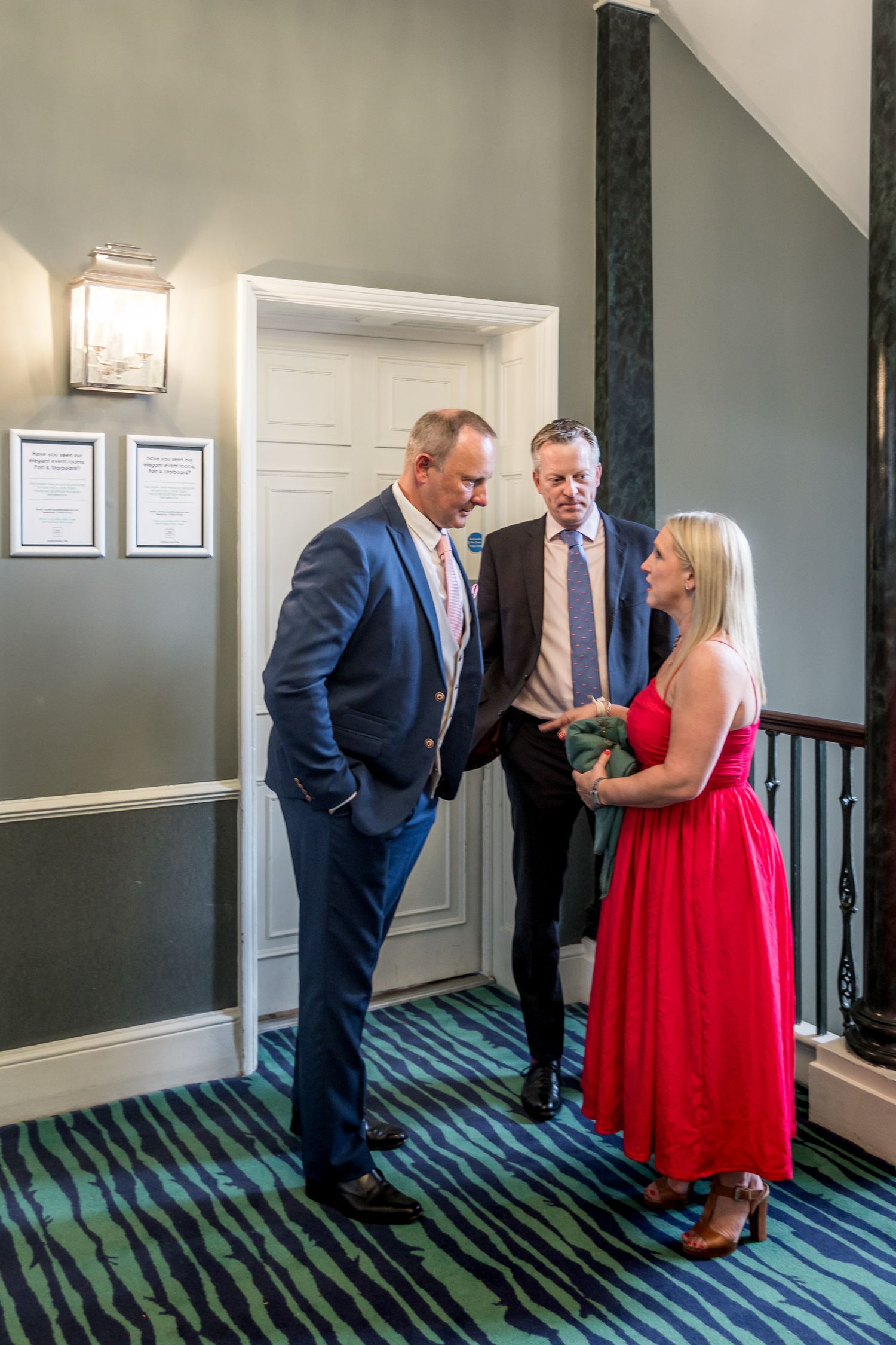 Guests in formal attire interact in hallway with blue-green patterned carpet.