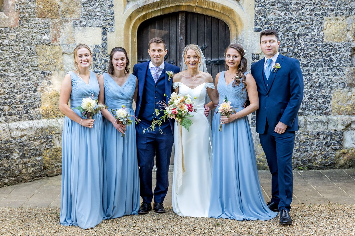 Wedding party poses in front of stone church wall wearing navy suits and light blue dresses.
