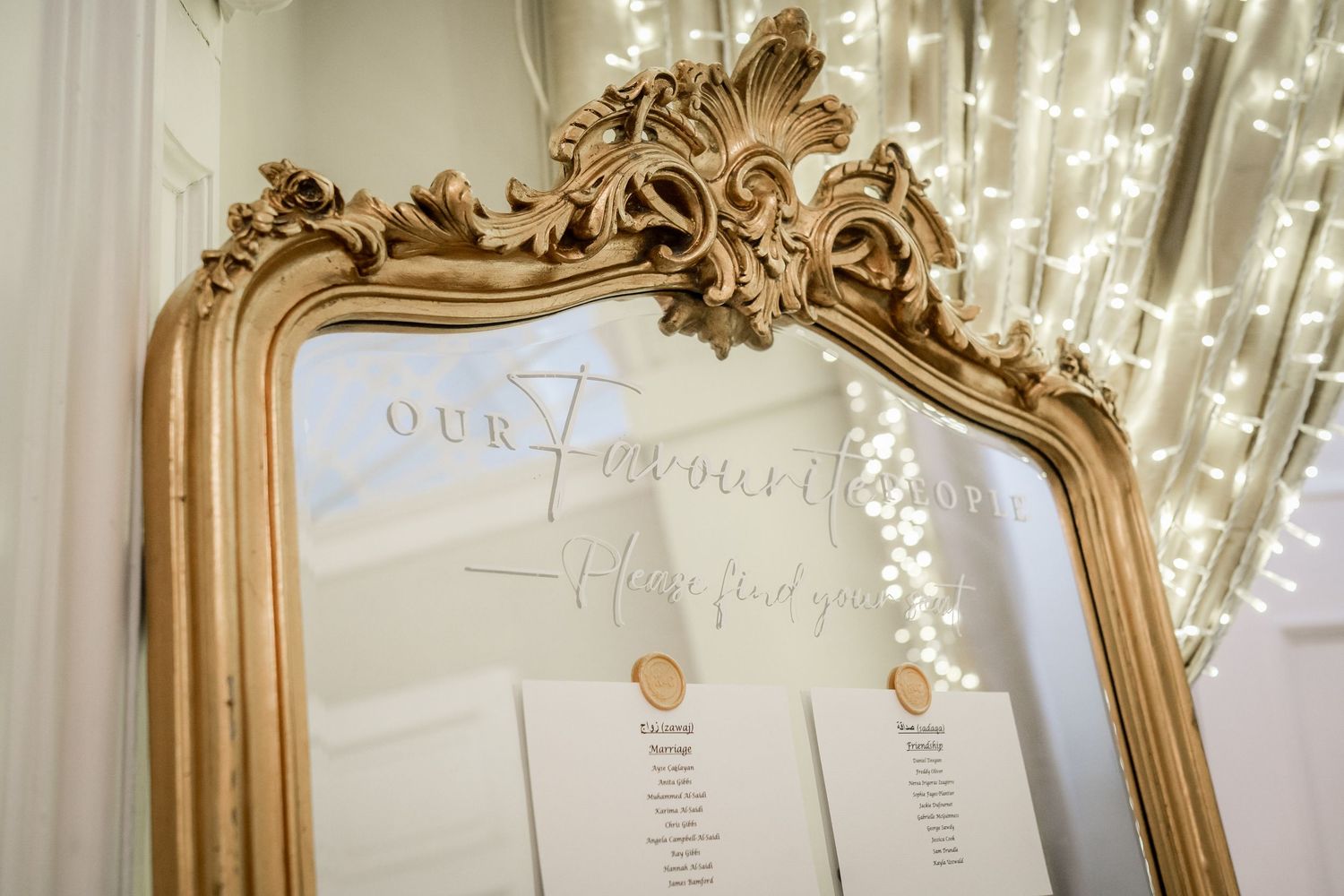 Ornate gold-framed seating chart display with string lights for wedding reception.