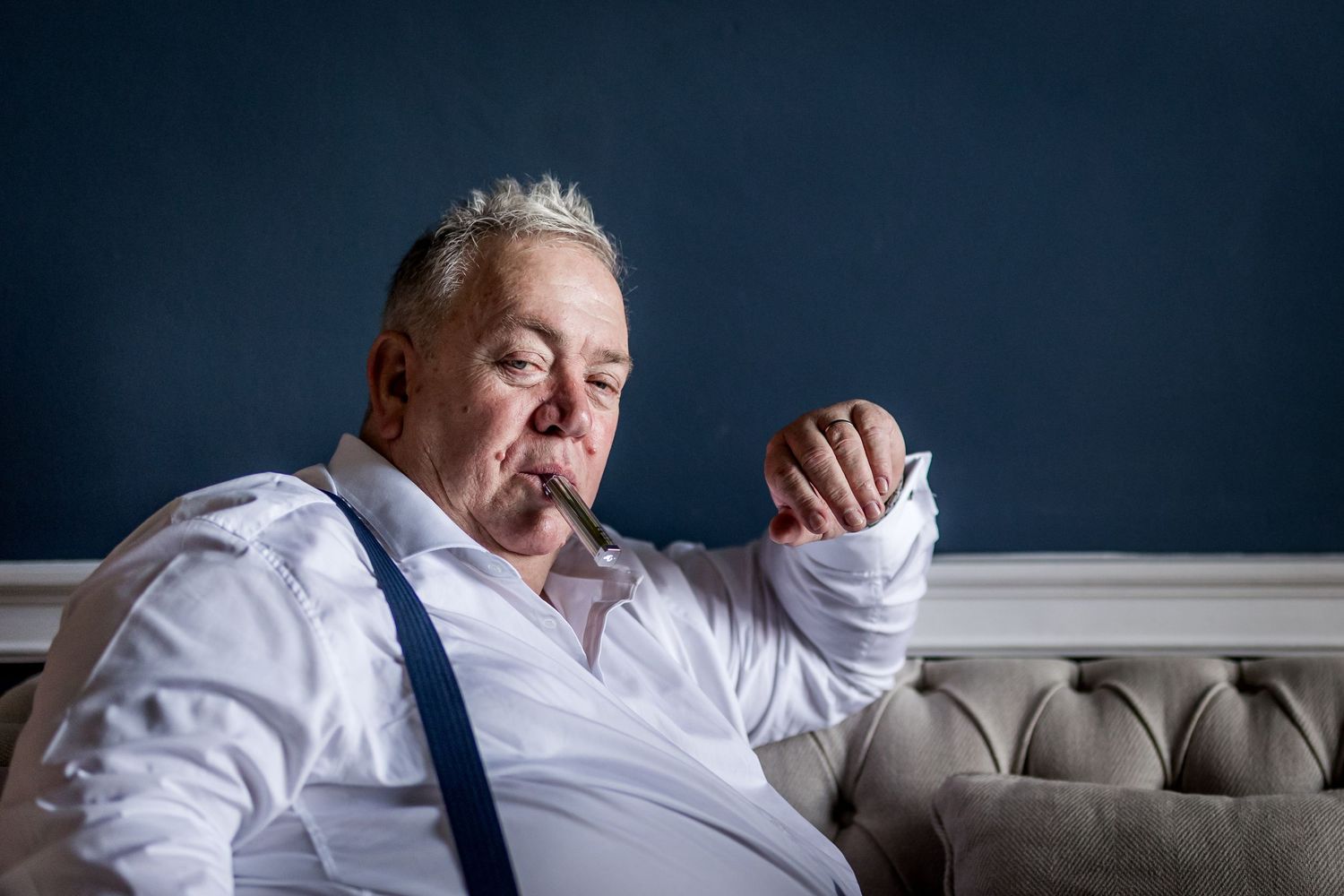Person in white shirt and suspenders reclining on tufted couch against blue wall.