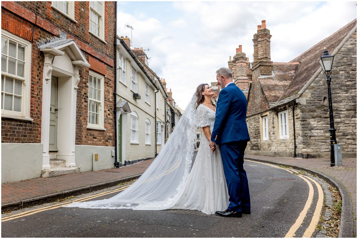 Newlyweds embrace on a curved street with classic British architecture in the background.