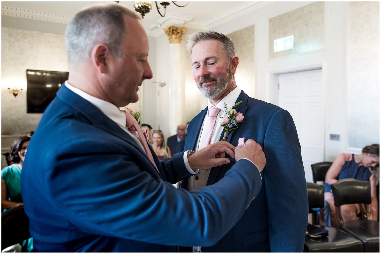Two people in blue suits adjusting boutonnieres and sharing a moment.