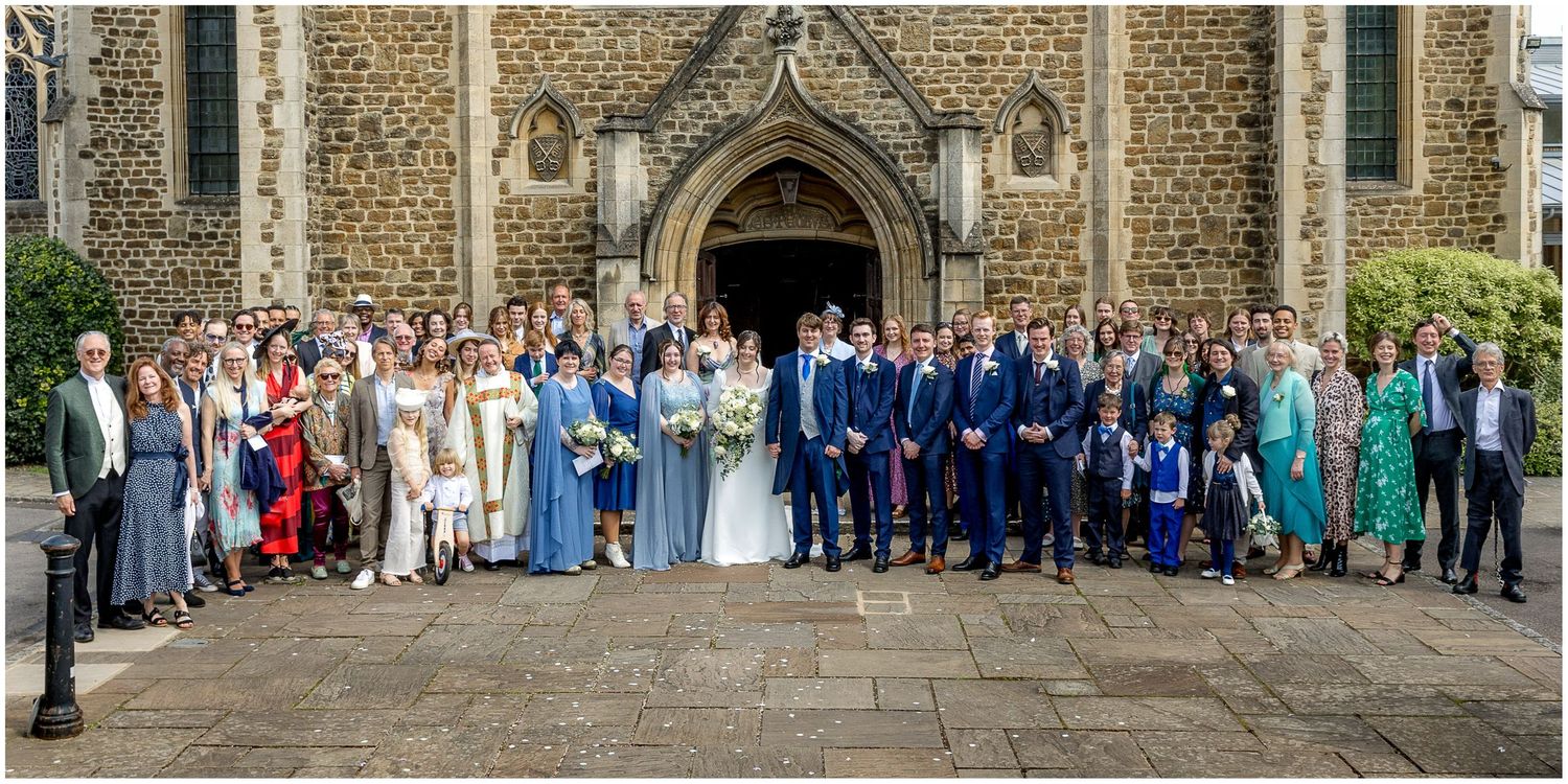Panoramic group photo of wedding party and guests in front of traditional English church.