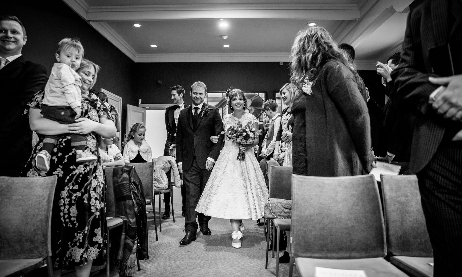 Black and white photo of wedding ceremony with guests seated in rows watching processional.