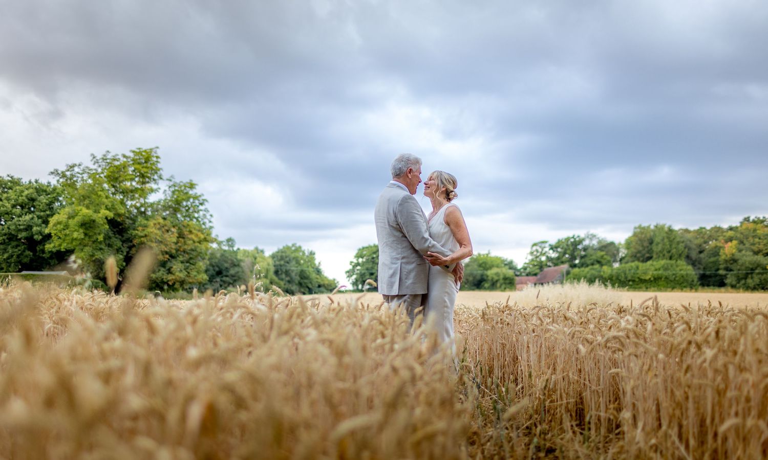 A couple stands together in a golden wheat field on a cloudy summer day with trees in the background.