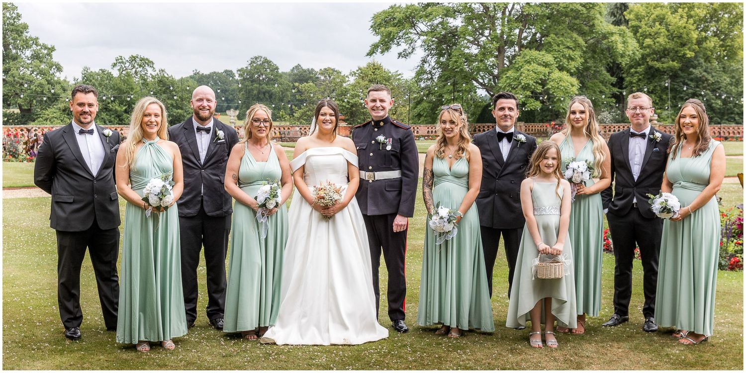 Wedding party in formal attire poses on green lawn with bridesmaids in sage dresses and groomsmen in black tuxedos.