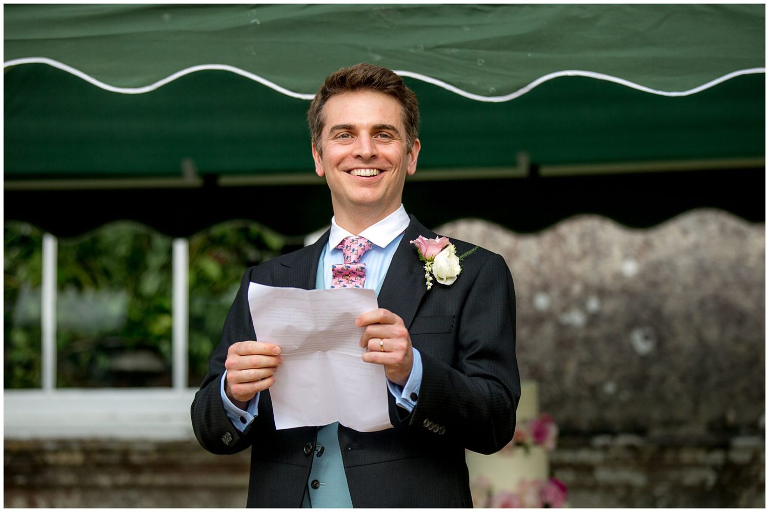 A person in a dark suit with a boutonniere smiles while reading from papers during an outdoor event under a green awning.