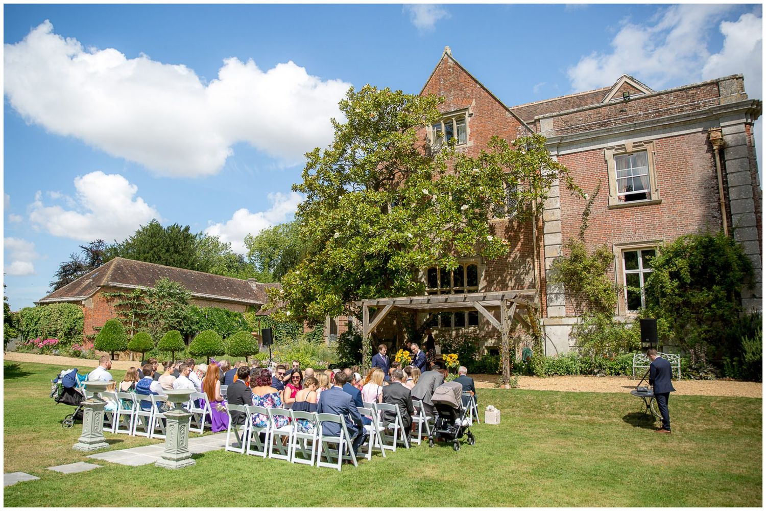 Panoramic view of an outdoor wedding ceremony setup with white chairs on a lush green lawn beside a historic brick building.