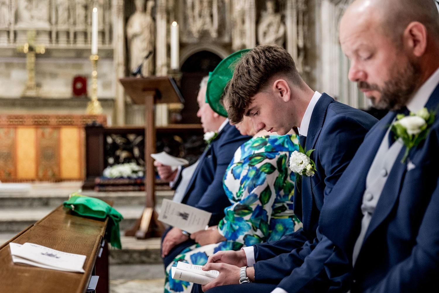 Wedding party members in navy suits seated in church pews during ceremony.