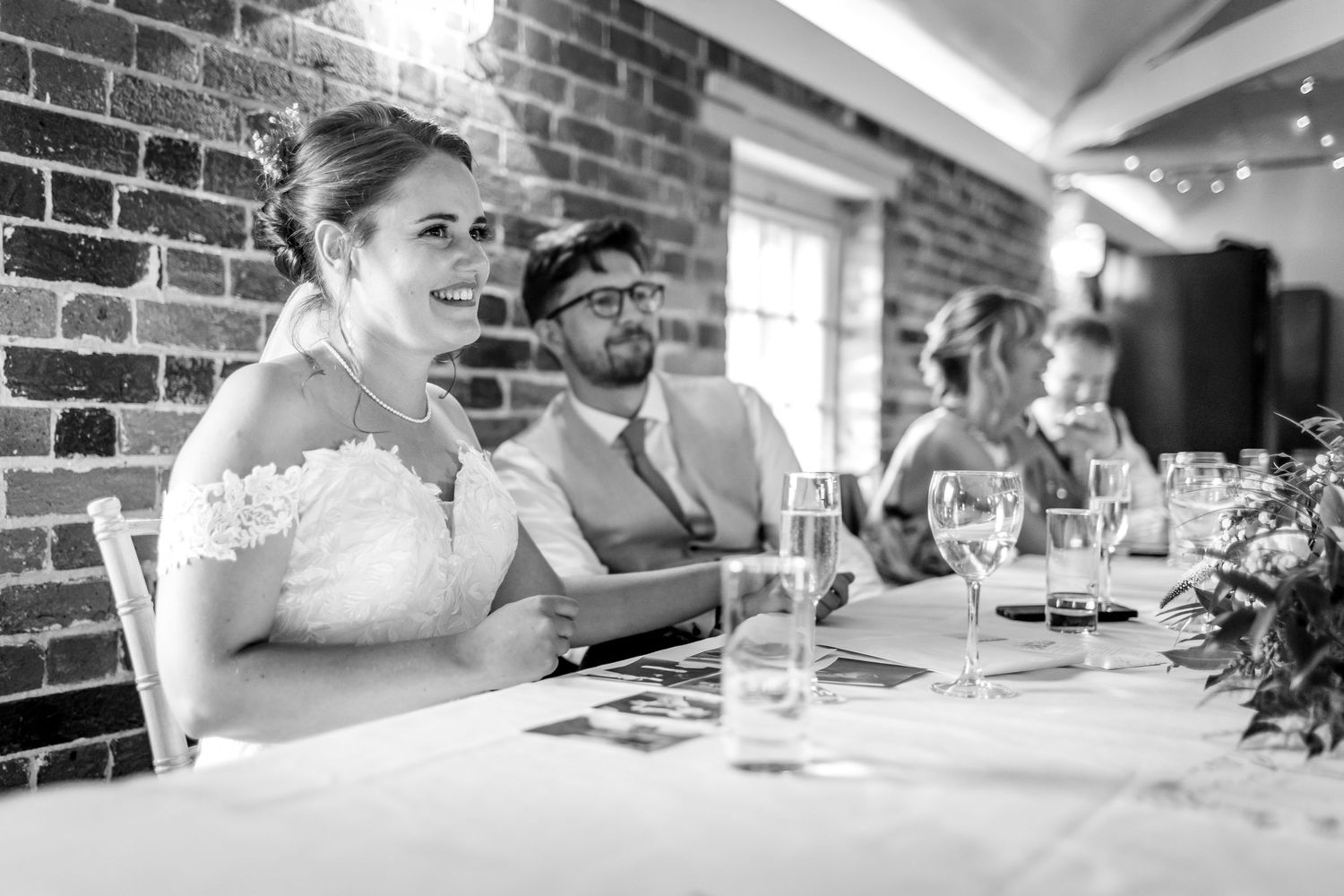 Black and white photo of guests dining at wedding reception with brick wall backdrop.