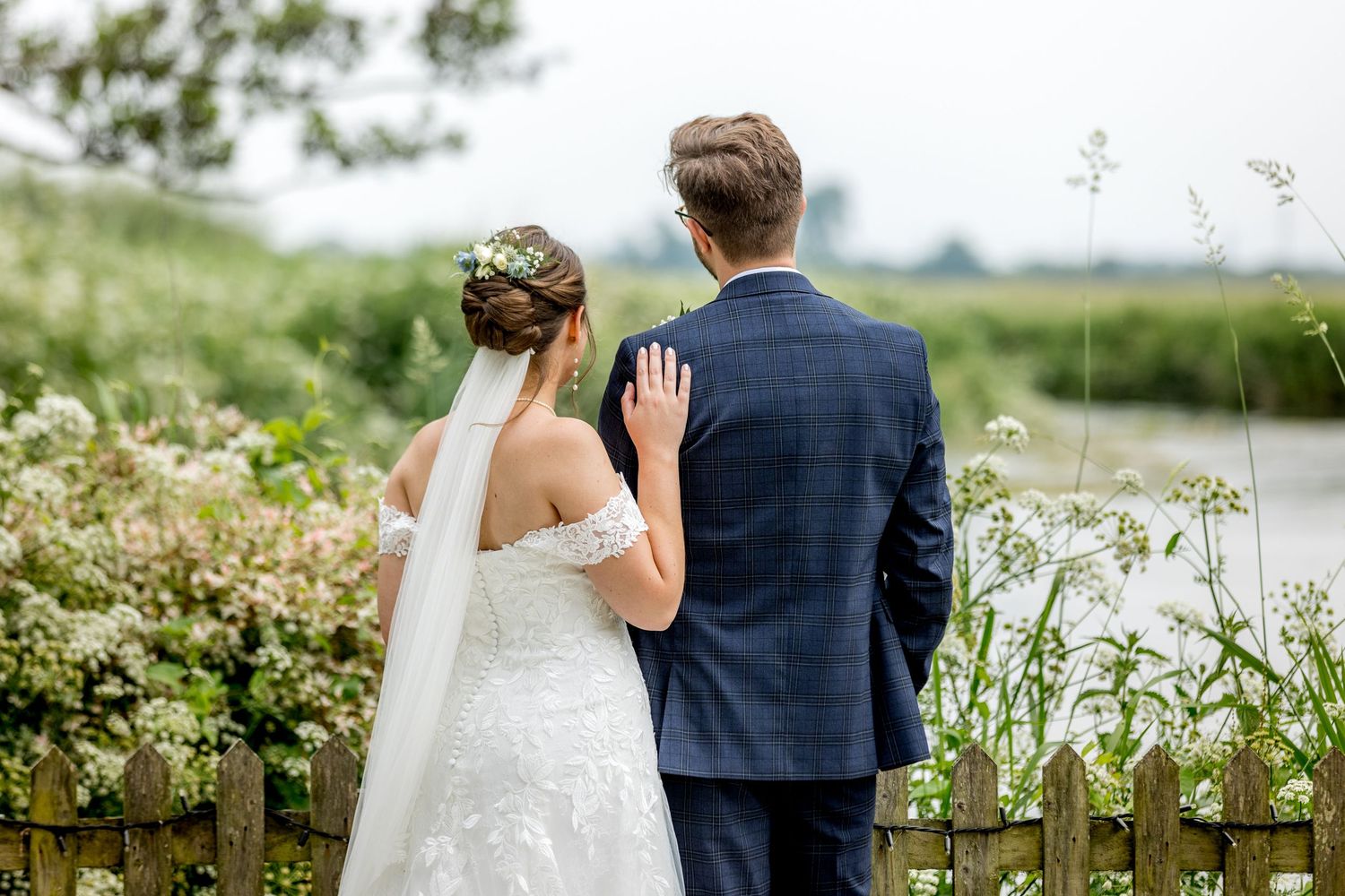 Wedding couple walks along rustic fence with wildflowers in countryside setting.