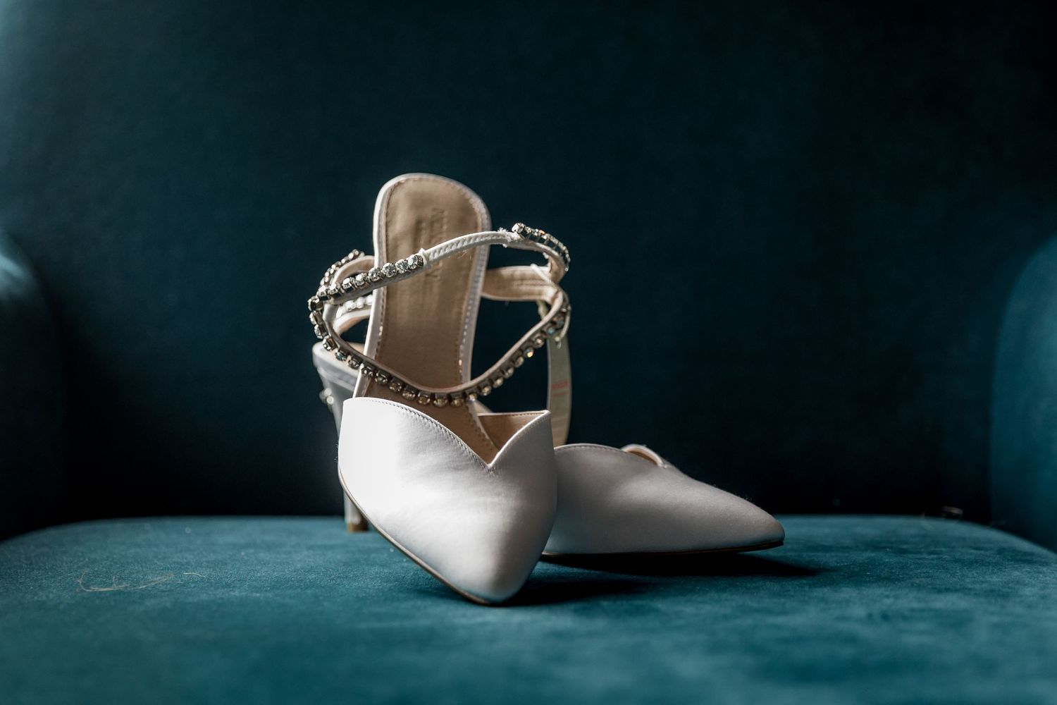 Elegant white bridal shoes displayed against a dark teal velvet background.
