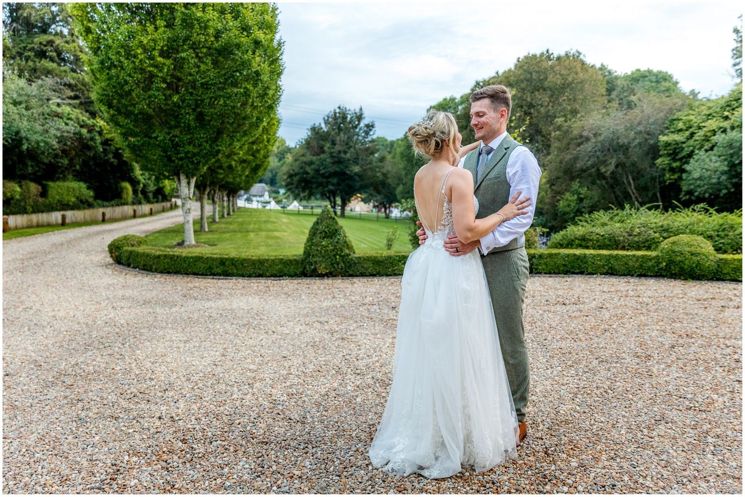 Newlyweds walk together on gravel path through manicured garden setting.