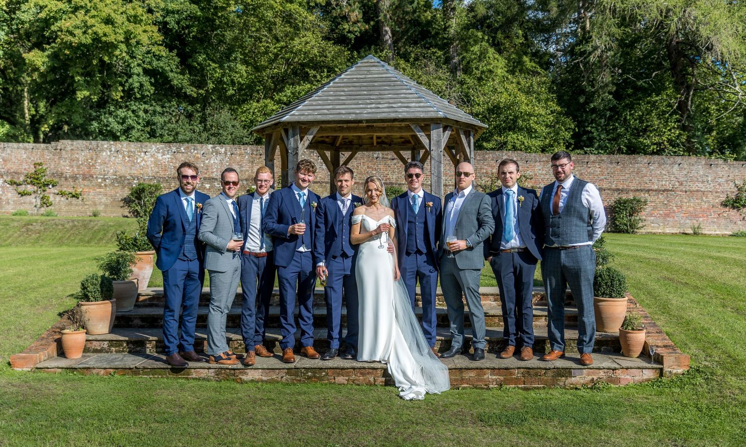 Wedding party poses on a lush green lawn in front of a rustic brick wall.