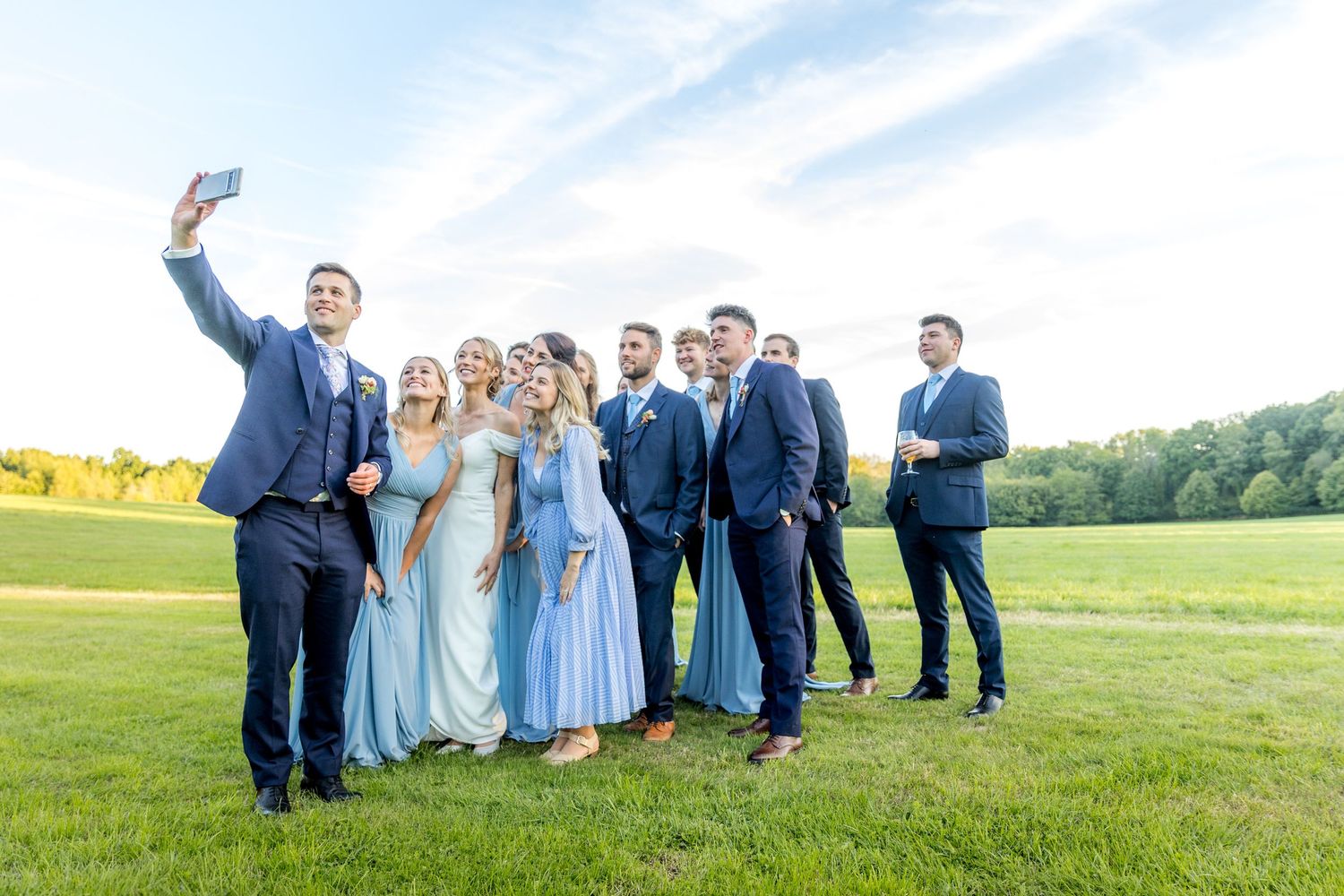 Wedding party members spread across green lawn taking group photos at outdoor venue.