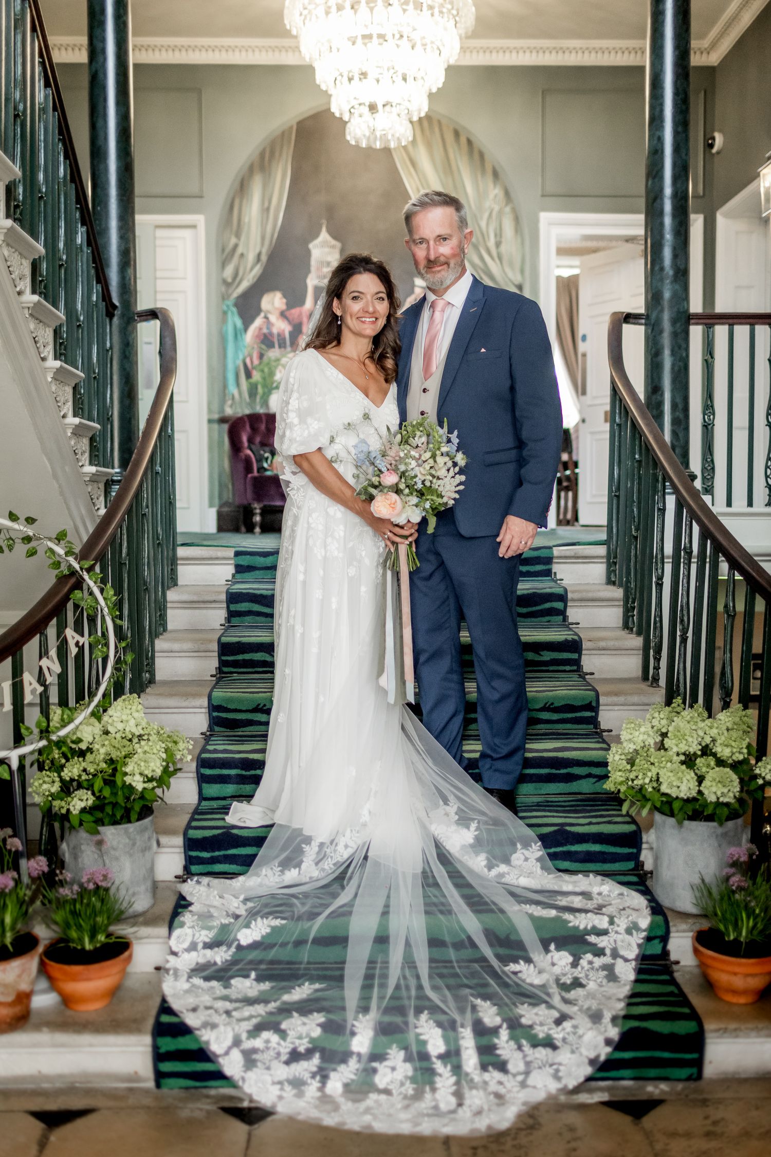 Elegant wedding photos on grand staircase with crystal chandeliers and ornate green railings.