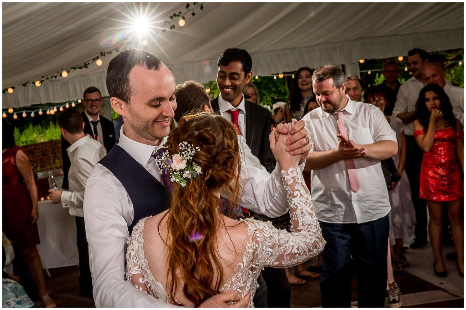 Couple dancing together at a wedding reception under a tent with guests watching and string lights overhead.