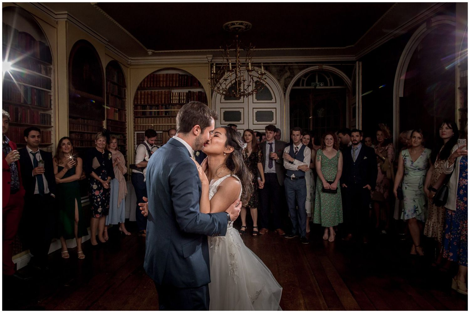 Newlyweds share their first dance surrounded by guests in an elegant ballroom with arched windows and chandelier lighting.