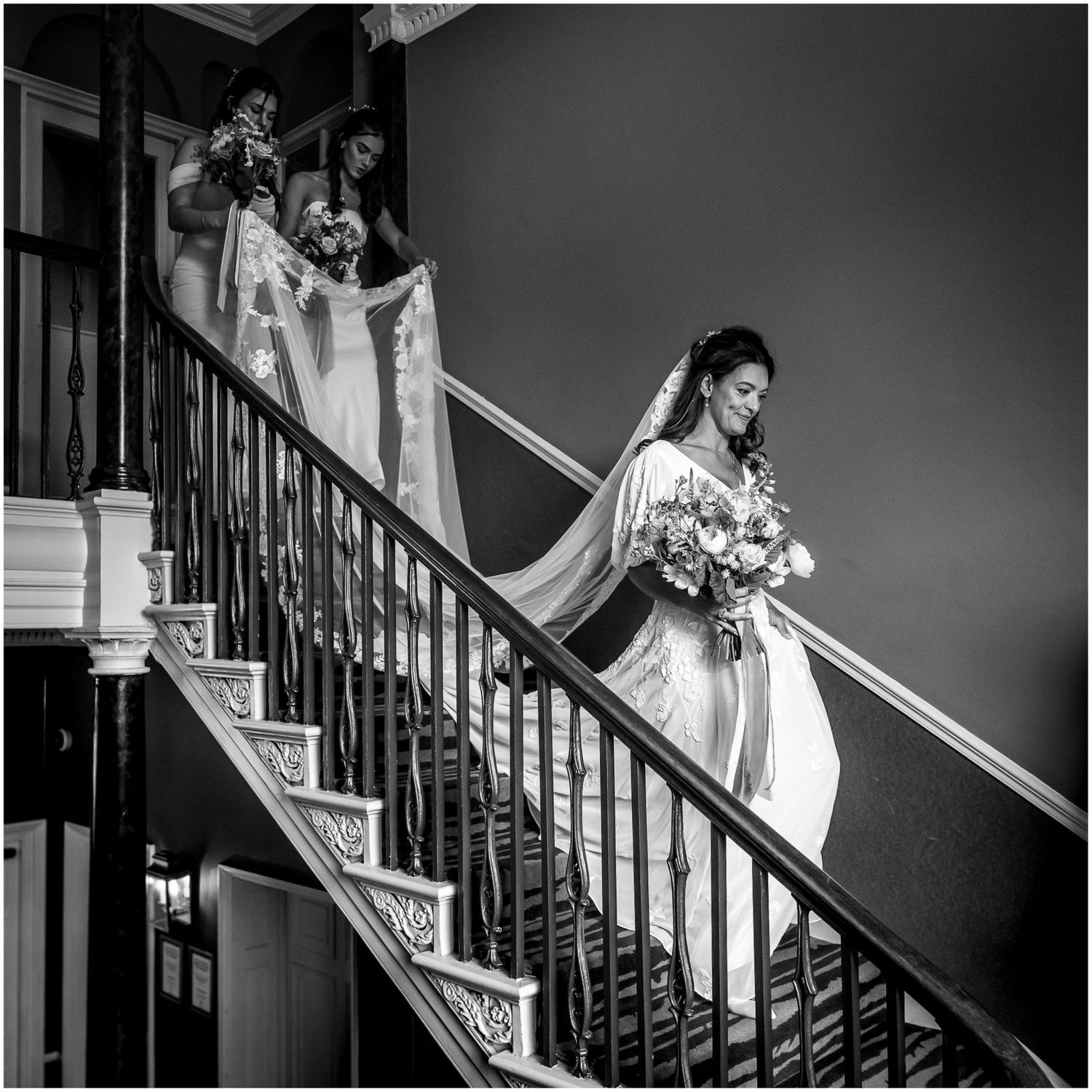 Black and white photo of person in wedding dress descending elegant staircase with bouquet.