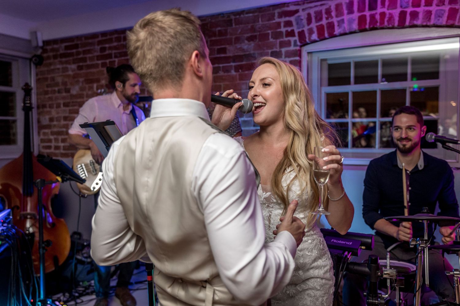 Couple performing together with live band at wedding reception in brick-walled venue with purple lighting.