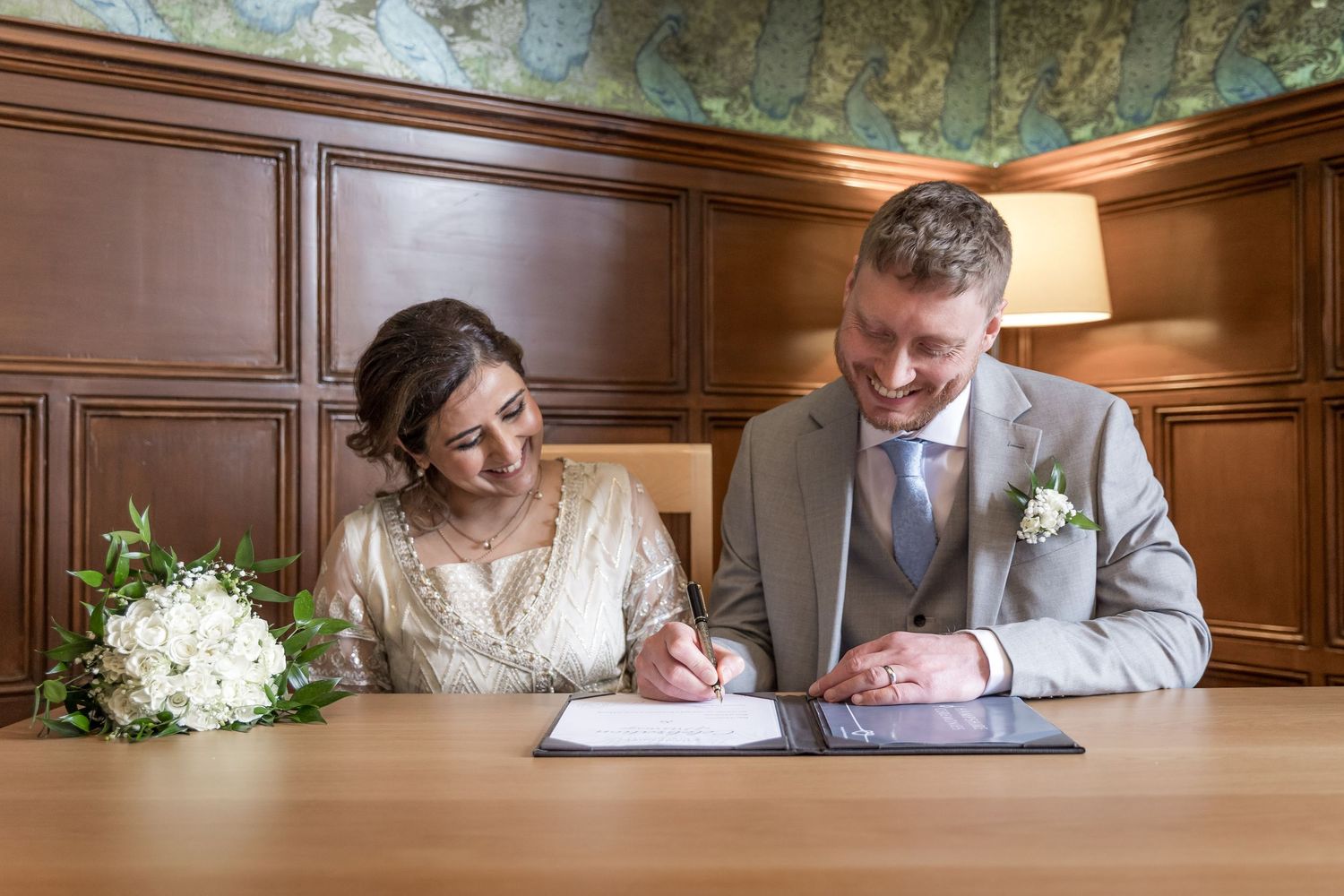 A couple signs documents at a wooden table with a white floral bouquet.