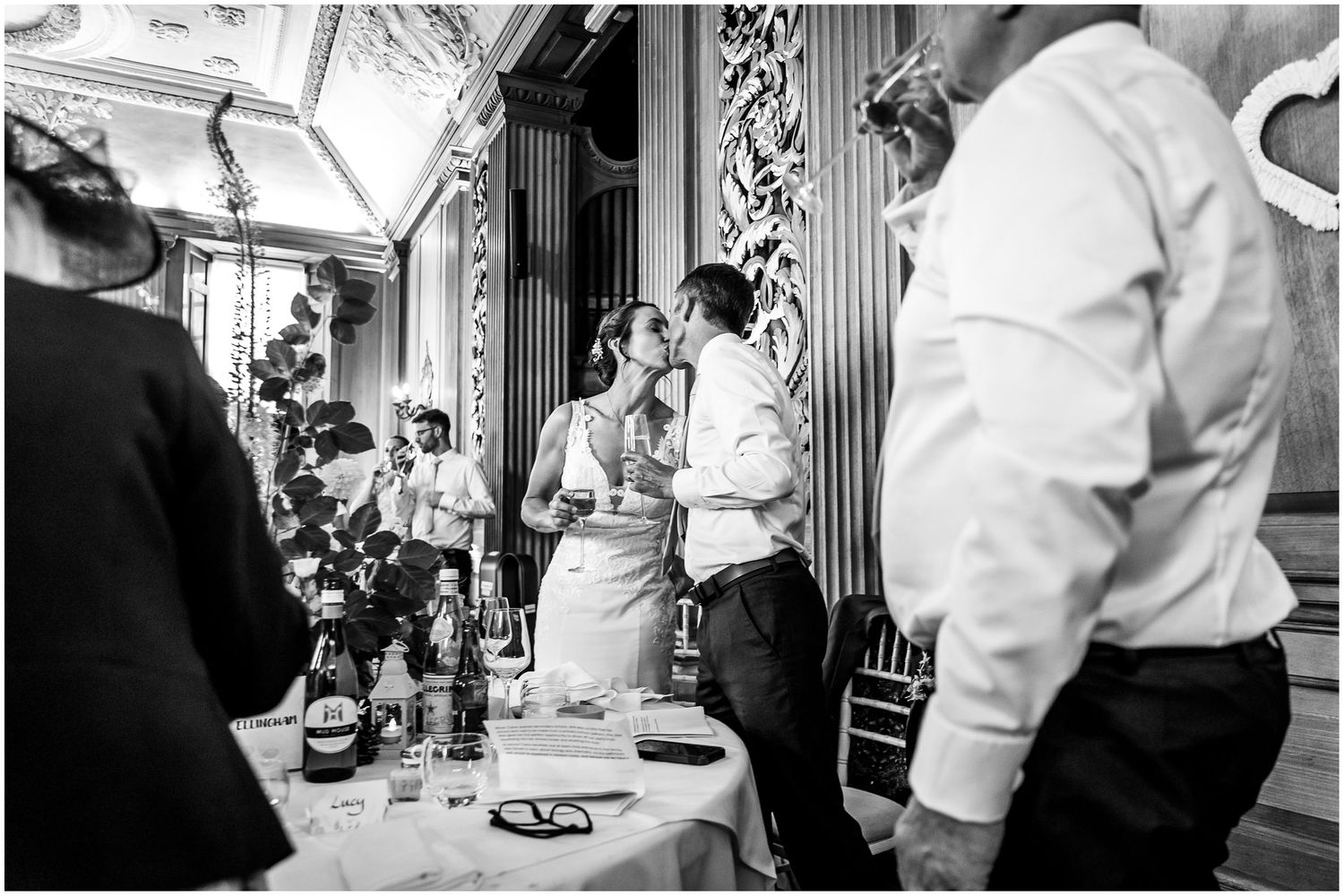 Black and white wedding reception photo of bride and groom sharing a kiss surrounded by guests in the hall at Walhampton in the New Forest