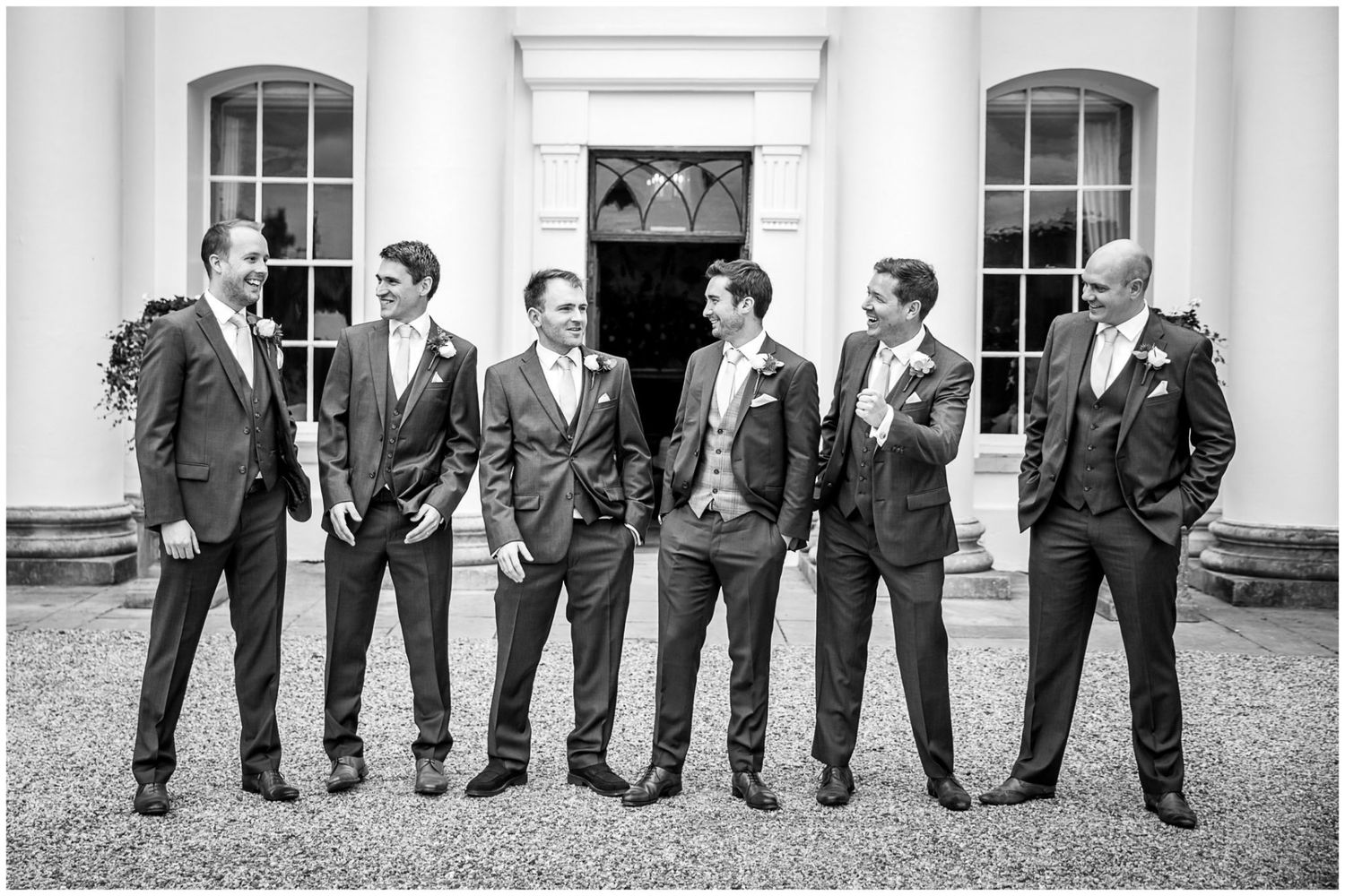 Wedding party in gray suits poses along a white columned building wall.