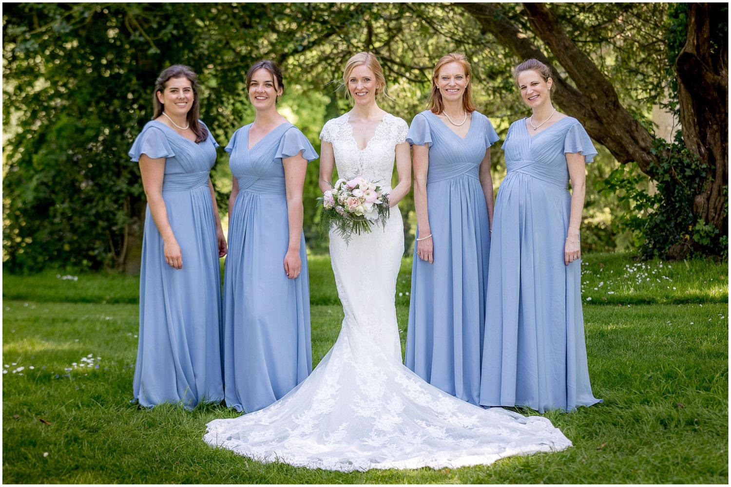 Bridesmaids in light blue dresses pose with bride in white gown on grassy lawn.