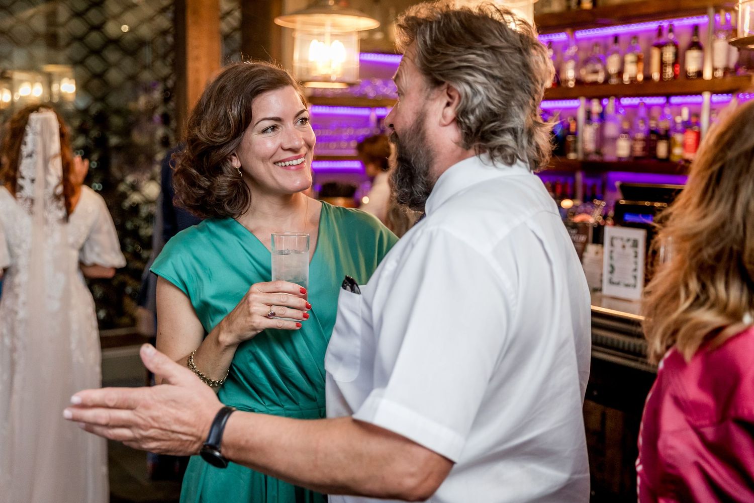 Partygoers engaged in lively conversation at a bar with purple ambient lighting.