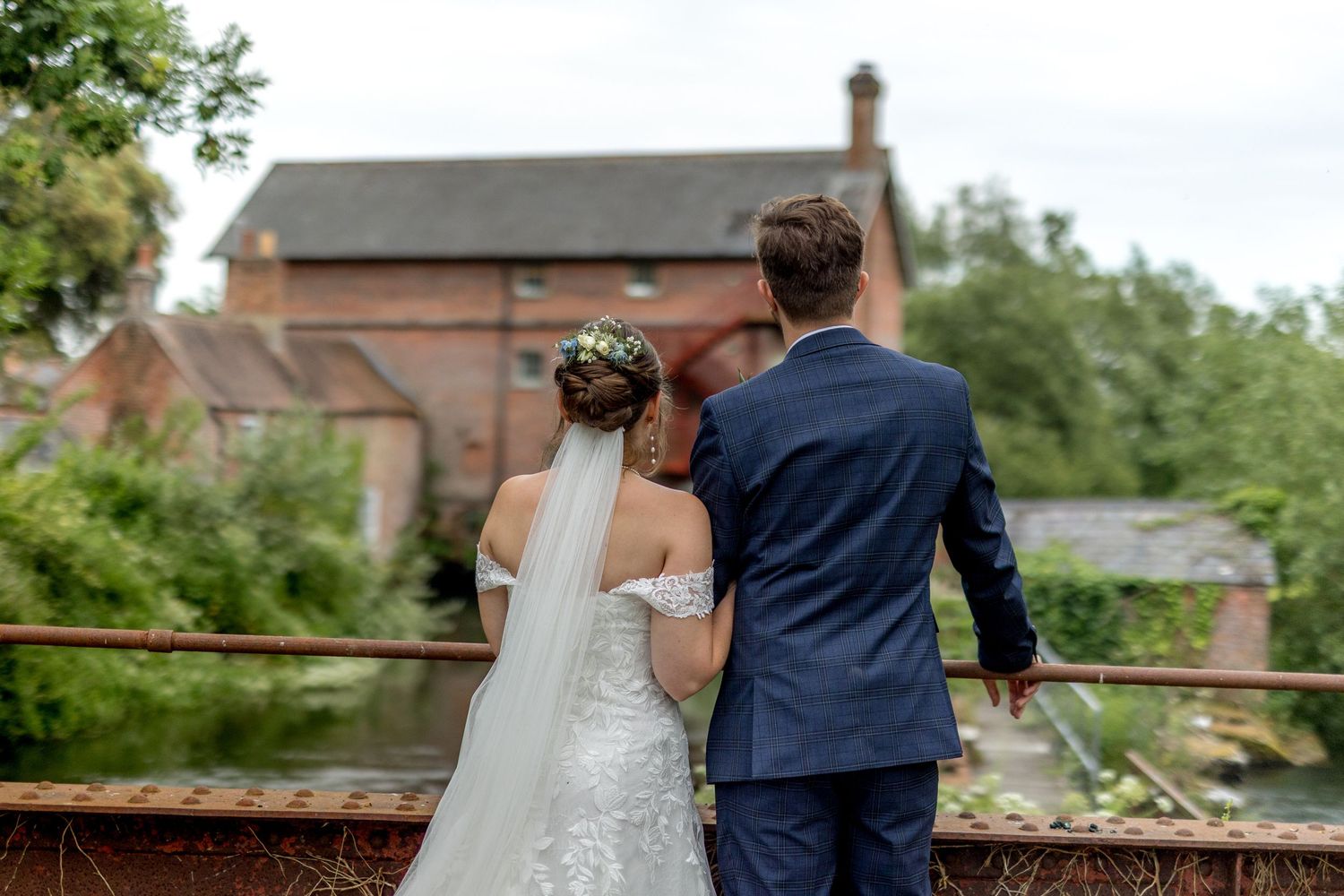 Wedding couple stands together on rustic bridge with brick buildings in background.