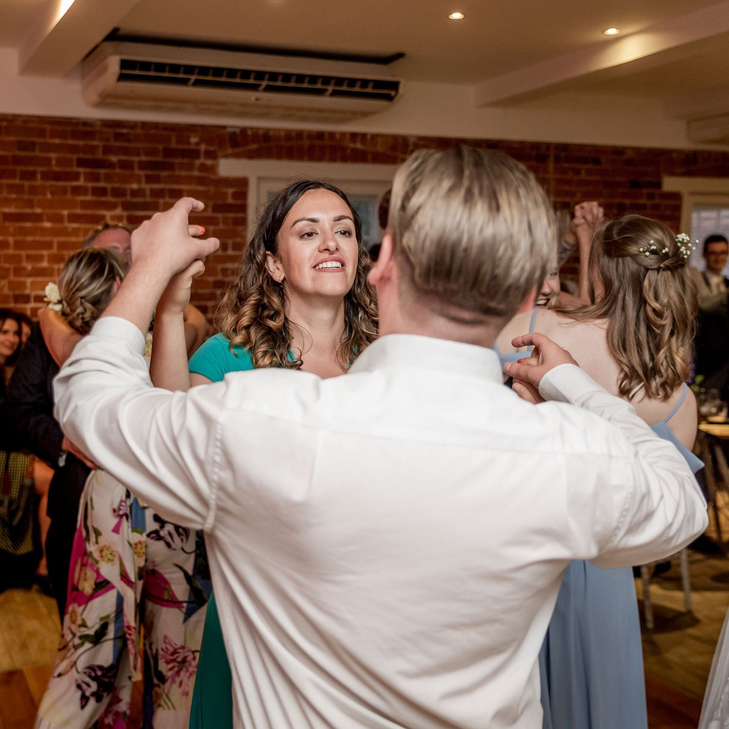 Wedding guests dancing with raised arms in a venue with exposed brick walls and overhead lighting.