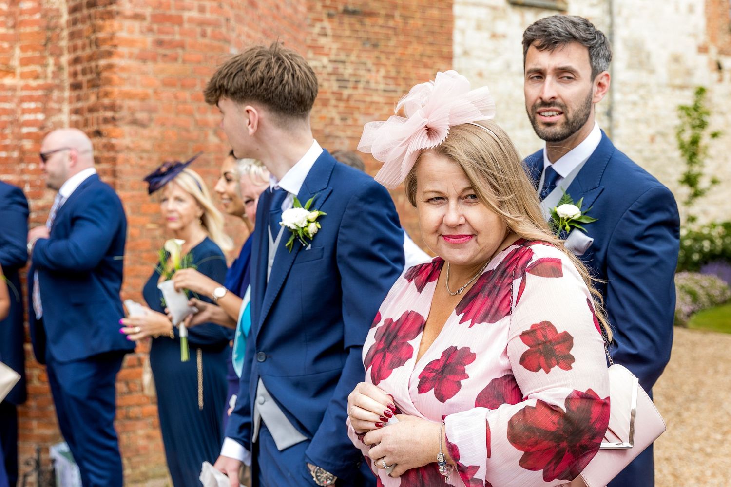 Wedding guests in formal attire gather outside brick venue walls.