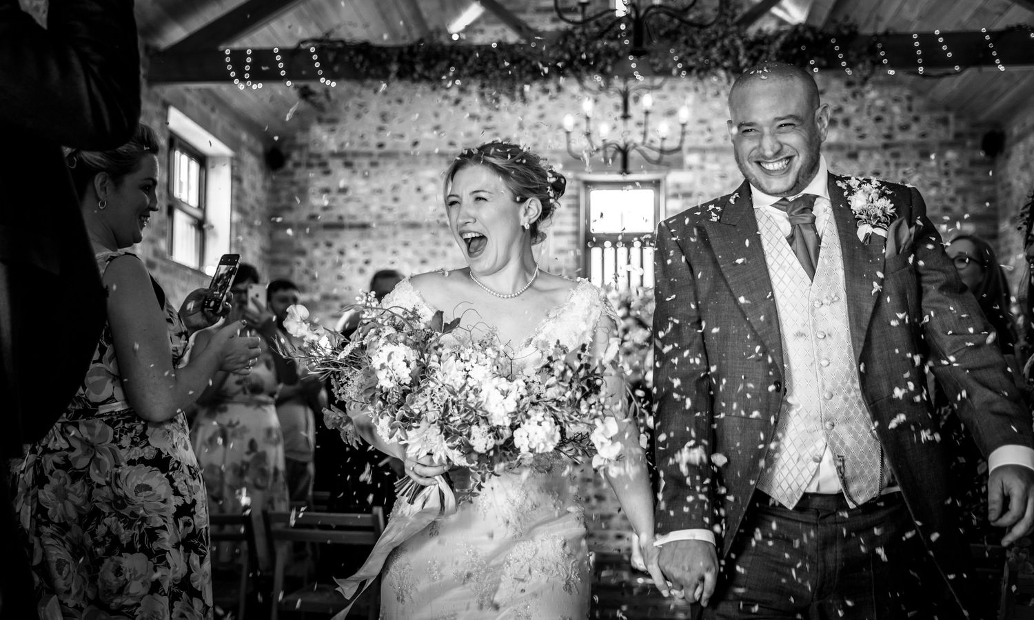 A joyful wedding couple walks down the aisle while guests shower them with confetti in a rustic stone venue.